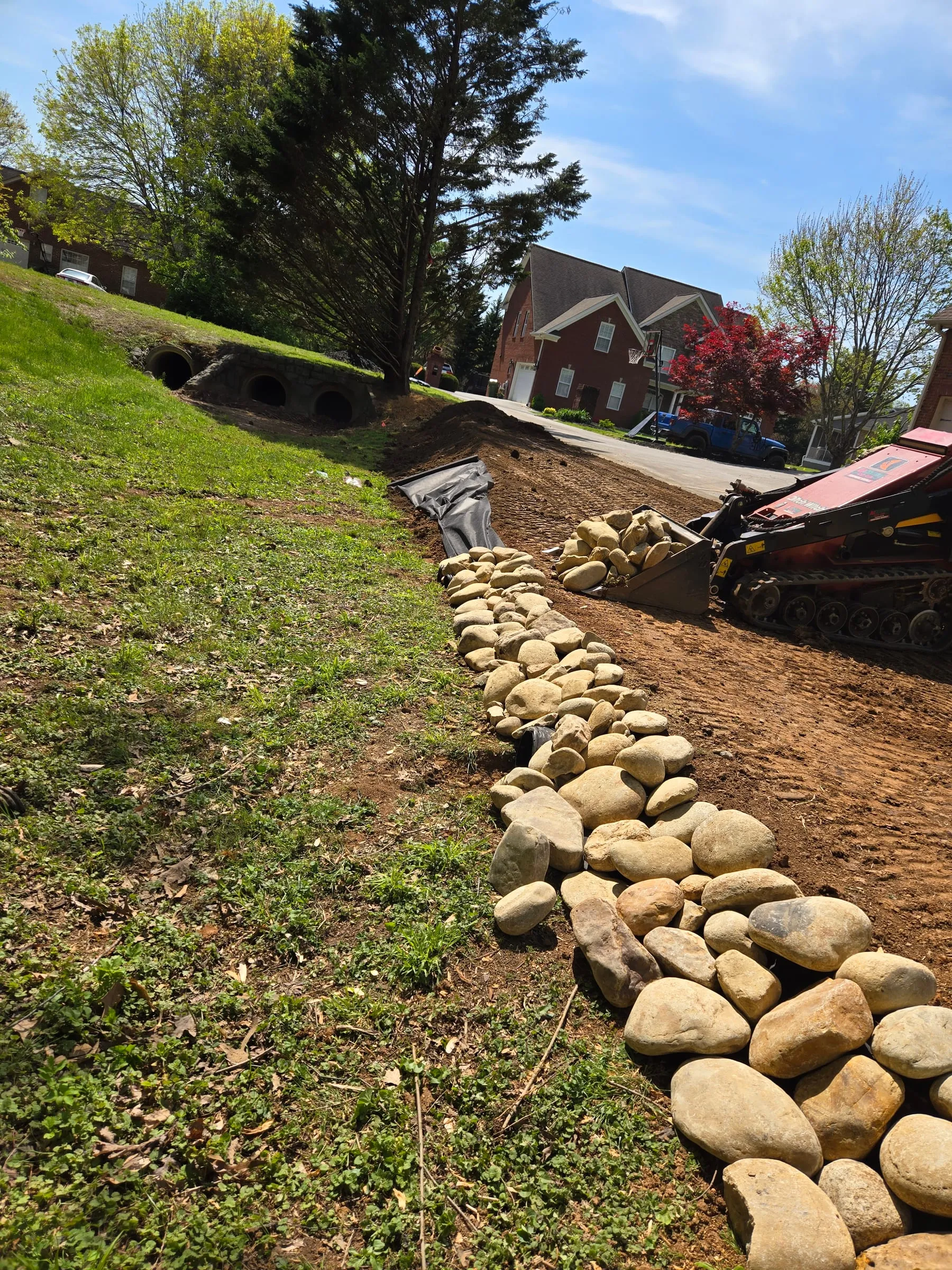 Compact track loader placing river rock over a fabric-lined drainage swale leading to culverts